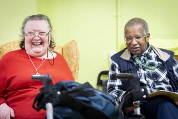 An older woman sitting in an arm chair and smiling. In front of her is a walking frame. An older man is sitting next to her wearing a sunflower lanyard.