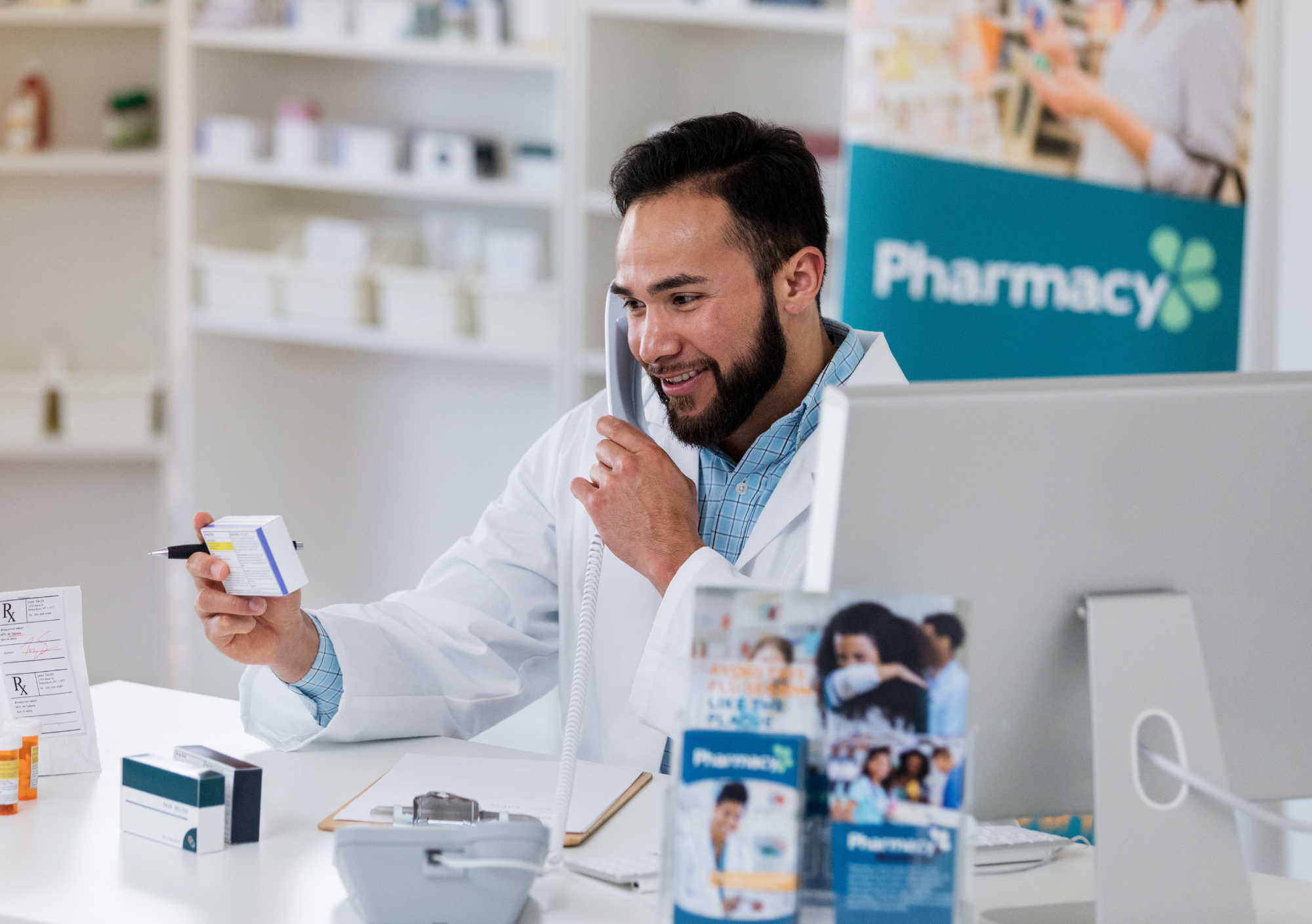 A pharmacist holding a box of medication in a pharmacy while on the phone with a customer.