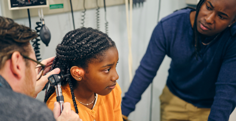 A doctor looking into the ear of a young girl in a doctor's office. The girl is accompanied by her father.