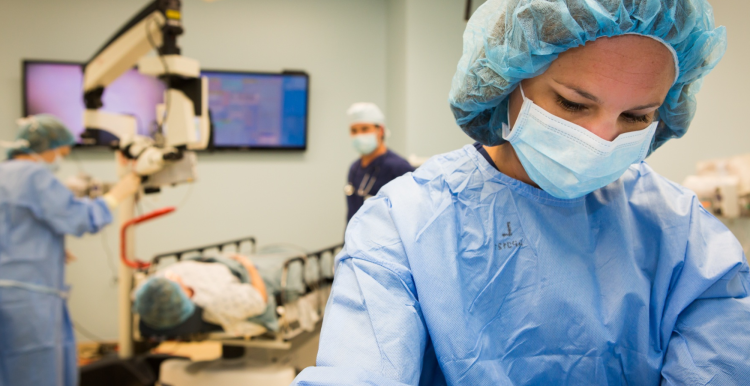 A medical professional wearing scrubs and a disposable face mask. In the background, a patient is lying down under a scanner with two more medical professionals watching over them.