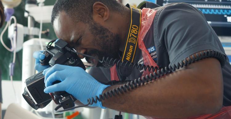 A Black medical professional taking a photo of something in a hospital setting. He is wearing blue disposable gloves and an apron.