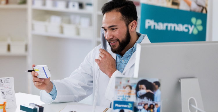 A pharmacist holding a box of medication in a pharmacy while on the phone with a customer.