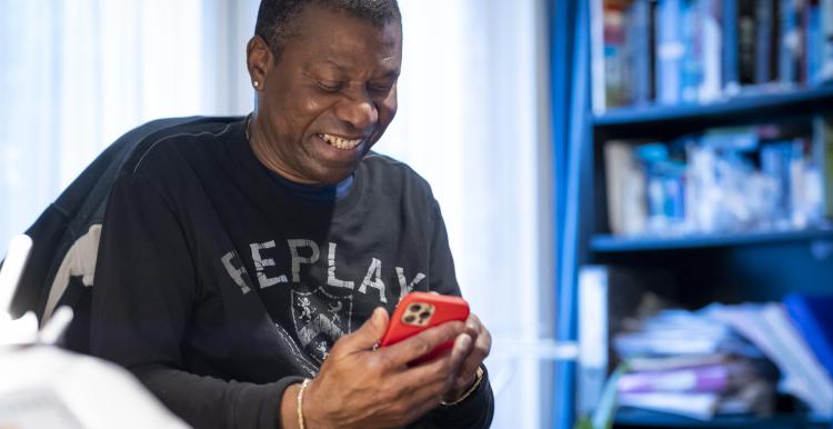A man sitting at a desk in front of a bookcase, looking down at a mobile phone.