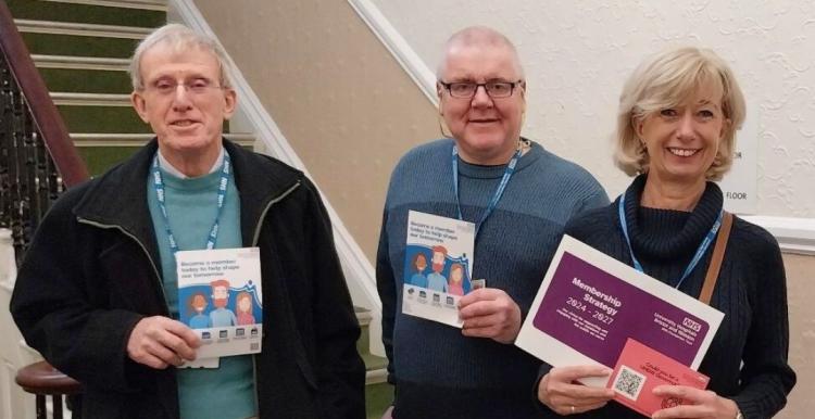 Three people wearing NHS lanyards, smiling and holding information booklets about becoming a UHBW governor.