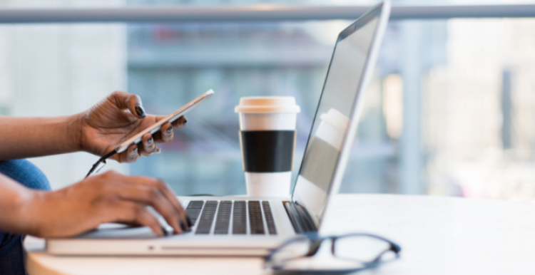 A woman's arms are visible, holding a phone. There is a laptop, coffee mug, and pair of glasses on a table.