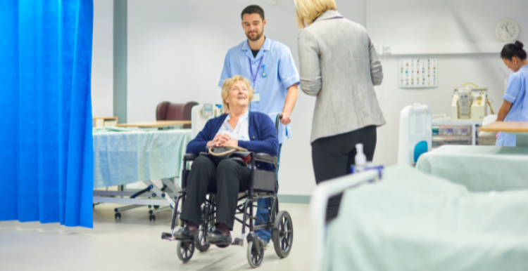 A person in a wheelchair being pushed by a member of hospital staff through a hospital ward. The person in a wheelchair is smiling at a person in a suit. Hospital beds and a blue curtain dividing the bays are visible on the ward.