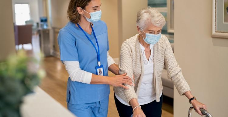 An older woman, using a walker and walking down a corridor. She is being supported by a nurse, who is holding her arm and wearing scrubs