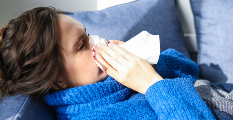 A woman in a blue jumper lying on a sofa and blowing her nose