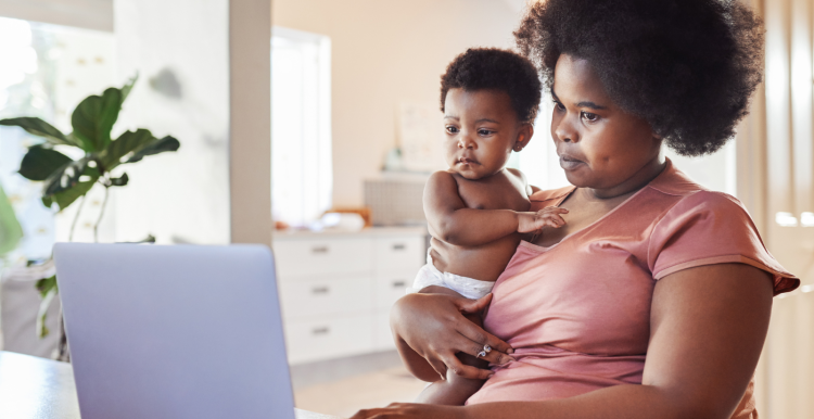 A Black mother holding her baby and looking at a laptop