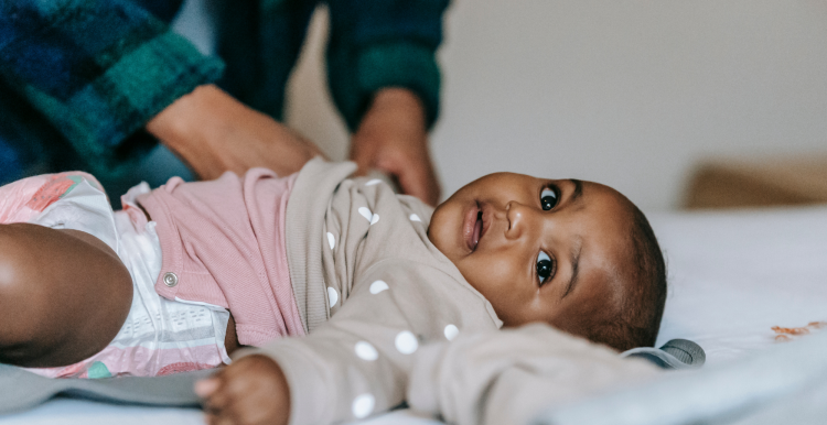 A young Black baby lying on a bed with a mother in the background