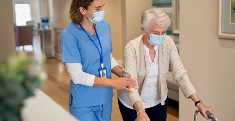 An older woman, using a walker and walking down a corridor. She is being supported by a nurse, who is holding her arm and wearing scrubs An older woman, using a walker and walking down a corridor. She is being supported by a nurse, who is holding her arm and wearing scrubs