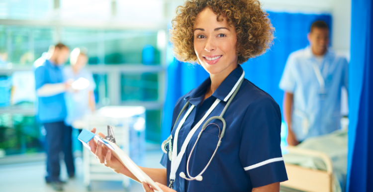 A medical professional standing in a hospital ward, holding a clipboard and wearing a stethoscope