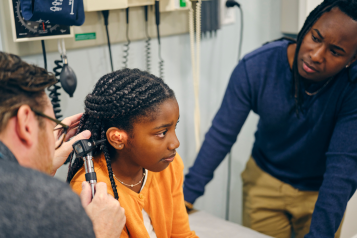 A doctor looking into the ear of a young girl in a doctor's office. The girl is accompanied by her father.