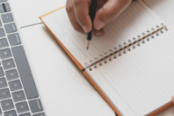 A person's hand writing in a notebook in front of a laptop.