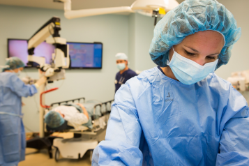 A medical professional wearing scrubs and a disposable face mask. In the background, a patient is lying down under a scanner with two more medical professionals watching over them.