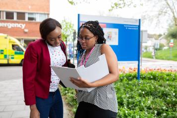 Two young women standing outside a hospital, looking at a clipboard.