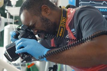 A Black medical professional taking a photo of something in a hospital setting. He is wearing blue disposable gloves and an apron.