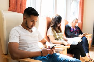 Three people sitting on a row of armchairs. Two people are looking at their mobile phones and the third is reading a leaflet.