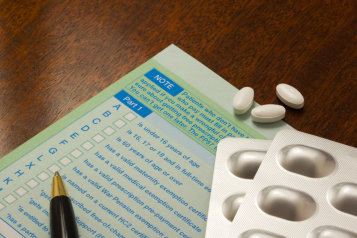 A green prescription form on a wooden surface. On top of the form is a blister pack of tablets, some loose tablets, and a pen.