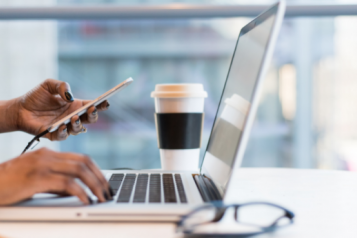 A woman's arms are visible, holding a phone. There is a laptop, coffee mug, and pair of glasses on a table.