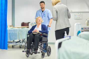 A person in a wheelchair being pushed by a member of hospital staff through a hospital ward. The person in a wheelchair is smiling at a person in a suit. Hospital beds and a blue curtain dividing the bays are visible on the ward.