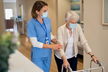 An older woman, using a walker and walking down a corridor. She is being supported by a nurse, who is holding her arm and wearing scrubs