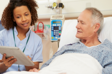 A man in a hospital bed, being shown something on a tablet by a member of hospital staff