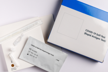 A blue and white box which says: COVID-19 Self Test (Rapid Antigen Test). Around the box is the extraction tube, swab, and test strip in a wrapper