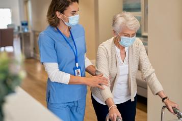 An older woman, using a walker and walking down a corridor. She is being supported by a nurse, who is holding her arm and wearing scrubs