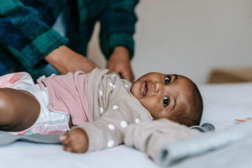 A young Black baby lying on a bed with a mother in the background