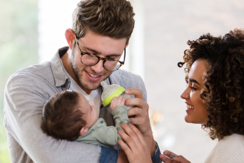 A father standing next to his wife, holding and feeding his baby