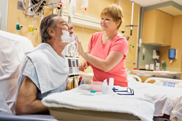 A woman helping a male patient in a hospital bed shave his facial hair