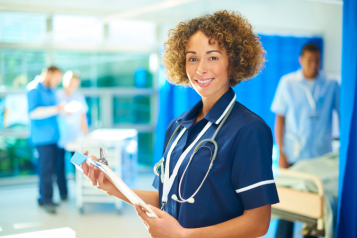 A medical professional standing in a hospital ward, holding a clipboard and wearing a stethoscope