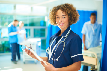 A medical professional standing in a hospital ward, holding a clipboard and wearing a stethoscope 