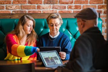 Two older people sitting at a table using tablets. A young person pointing at something on one of the tablets.