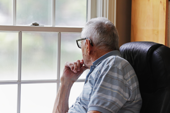 An older man is sitting quietly with his hand on his chin, looking out the window.