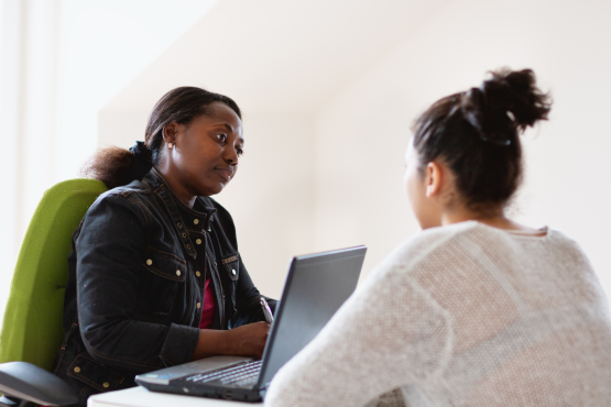 A woman sitting behind a laptop, looking thoughtful. She is talking to another woman who is sitting across from her.