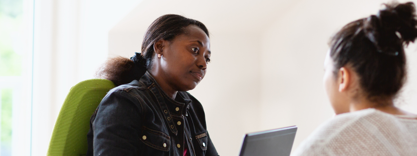 A woman sitting behind a laptop, looking thoughtful. She is talking to another woman who is sitting across from her.