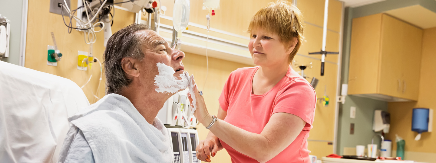 A woman helping a male patient in a hospital bed shave his facial hair