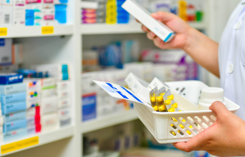 Close up showing a pharmacist's hands and arms. They are filling a prescription basket, standing in front of shelves containing medication boxes.