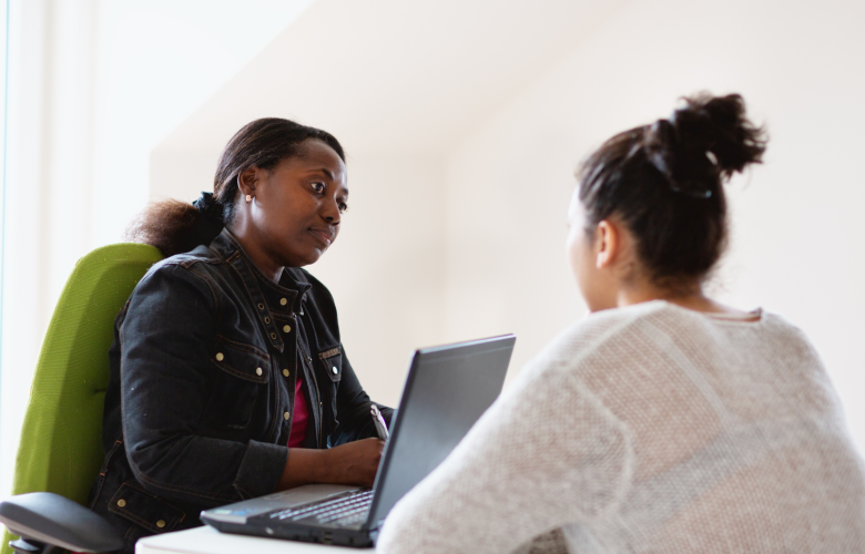A woman sitting behind a laptop, looking thoughtful. She is talking to another woman who is sitting across from her.