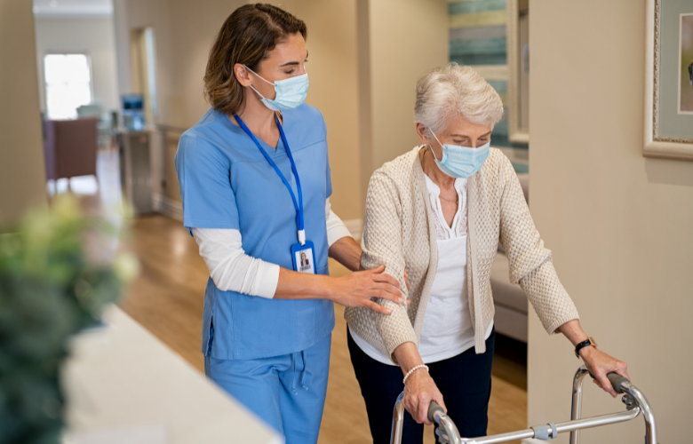 An older woman, using a walker and walking down a corridor. She is being supported by a nurse, who is holding her arm and wearing scrubs