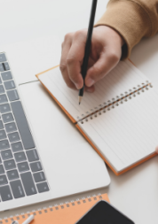 Close-up of a person's hand writing in a notepad, in front of a laptop.