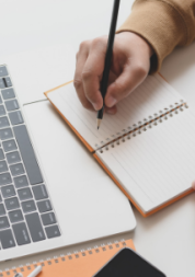 A person writing in a notebook in front of a laptop