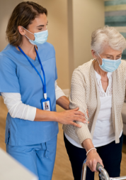 An older woman, using a walker and walking down a corridor. She is being supported by a nurse, who is holding her arm and wearing scrubs